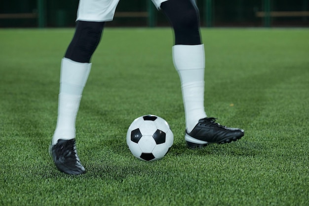 Piernas de jugador de fútbol en calcetines blancos y calzado deportivo negro haciendo ejercicio con balón de fútbol sobre césped verde en el estadio
