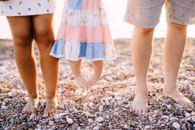 Piernas de mamá papá e hija pequeña en un vestido de pie en una playa de guijarros