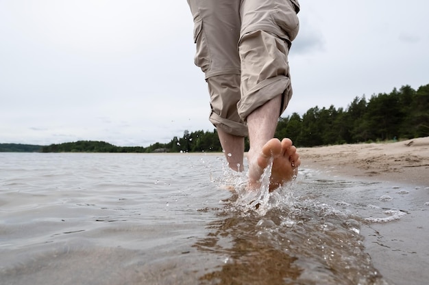 Piernas masculinas descalzas en pantalones enrollados caminan sobre el agua a lo largo de la orilla arenosa