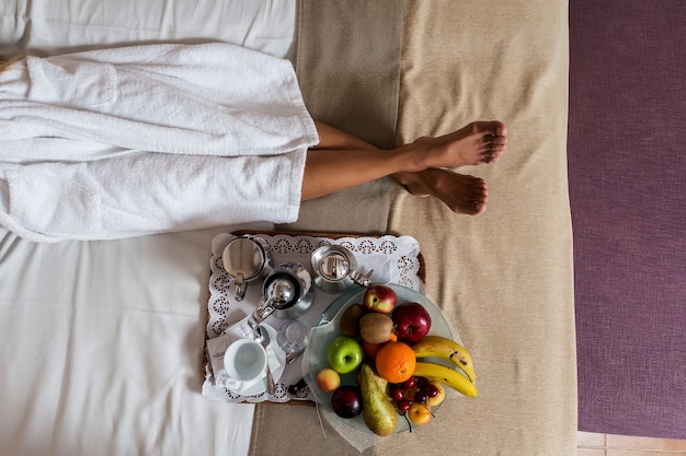 Piernas de mujer acostada en la cama con plato de frutas