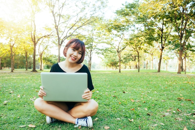 Las piernas de la mujer asiática joven en la hierba verde con la computadora portátil abierta