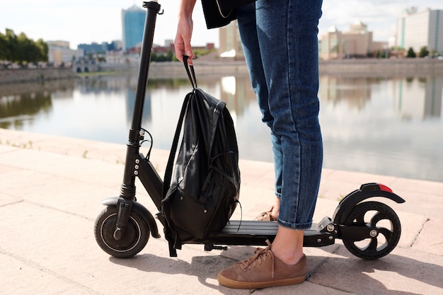 Piernas de mujer joven en jeans sosteniendo una mochila de cuero negro sobre scooter eléctrico mientras está de pie contra la orilla del río y el paisaje urbano