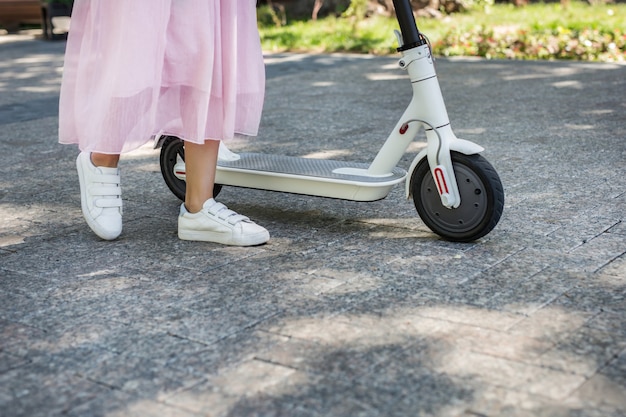 Piernas de la mujer que van cerca de scooter eléctrico ecológico en el asfalto de la ciudad en un día soleado de verano