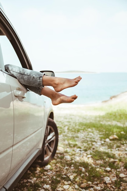 Las piernas de la mujer sobresalen de la playa del mar de la ventana del coche en las vacaciones de verano de fondo