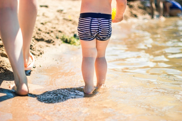 Piernas de niño pequeño y su madre caminando por la orilla del mar en una playa.