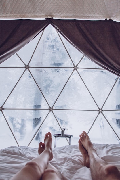 Piernas de una pareja de hombre y mujer en la cama contra el fondo de un bosque cubierto de nieve en un campamento de cúpula Concepto de estilo de vida de vacaciones Glamping