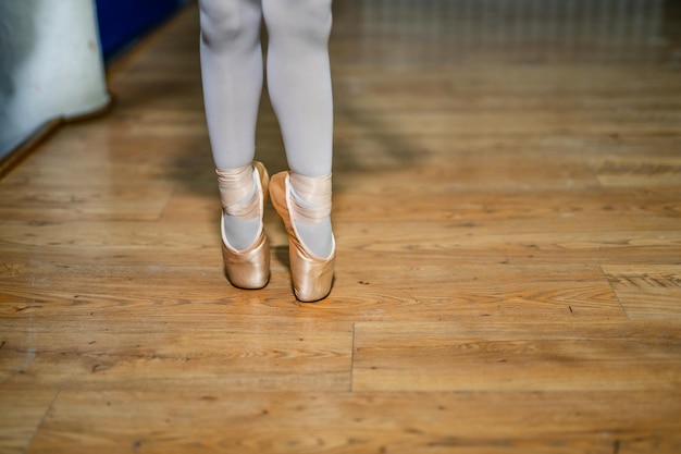 Piernas de una pequeña bailarina en zapatillas de ballet doradas y calcetines blancos posando de puntillas en la sala de ballet en el suelo Primer plano