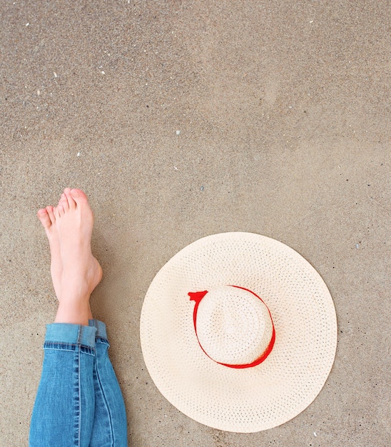 piernas y pies de mujer caucásica en jeans sentada junto al mar en la costa con sombrero cerca de la arena