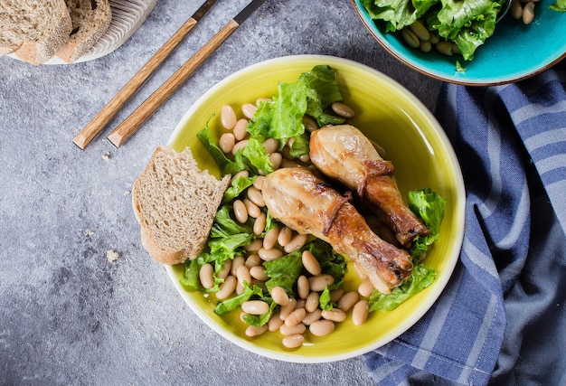 Piernas de pollo al horno con frijoles blancos y hojas de lechuga en un plato sobre fondo de mesa de piedra gris