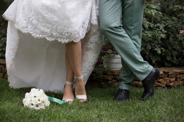Las piernas de los recién casados están sobre la hierba verde y el ramo de la boda.