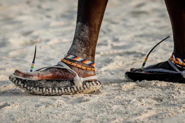 Piernas tribales masai con un colorido brazalete y sandalias hechas de neumáticos de automóvil en la playa de arena cerca de la isla de Zanzíbar, Tanzania, África Oriental