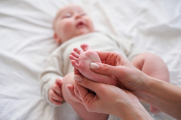 Pies de bebé en manos de la madre. La mujer caucásica joven hace el masaje para el bebé infantil feliz en la cama blanca en casa. Cuidado del bebé, deporte y feliz maternidad.