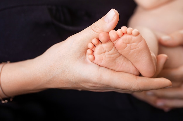 Pies del bebé recién nacido en manos de la madre en escena negra. Mamá sosteniendo a su hijo.