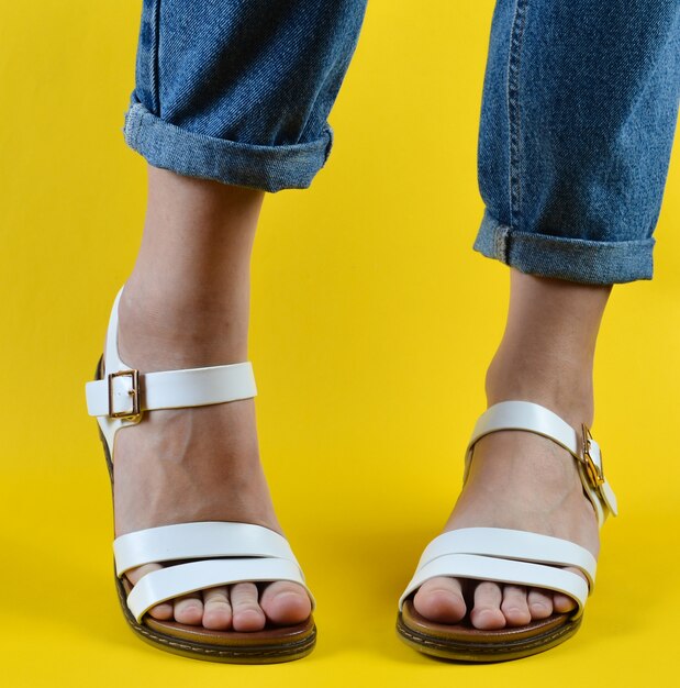 Pies femeninos vestidos con jeans y calzados con sandalias posando en una pared amarilla. Moda femenina. Calzado de verano para mujer