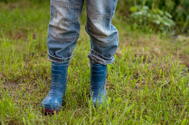 Los pies de los niños en jeans y botas de goma azul sucias de pie sobre la hierba