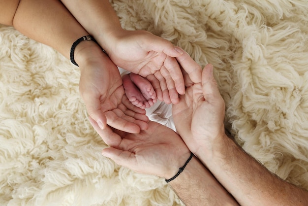 Pies del recién nacido en las palmas de los padres Las palmas del padre y la madre sostienen el pie del bebé recién nacido en una manta blanca flokati Fotografía de los dedos de los pies de un niño, talones y pies