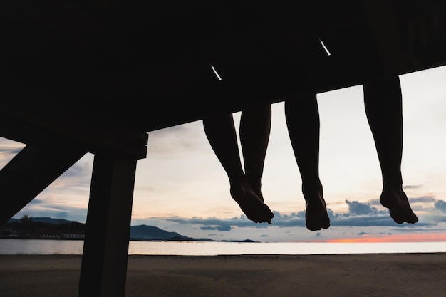 Pies de silueta de pareja sentada en el muelle en la playa al atardecer