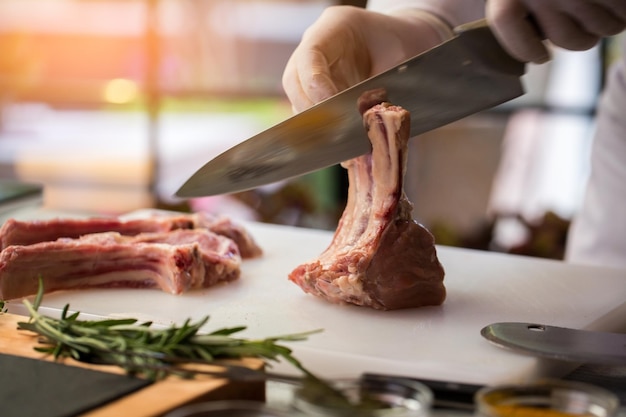 Pieza de carne cruda con huesos. La mano sostiene un cuchillo de cocina. Costillas de ternera y romero. Córtalo en trozos iguales.