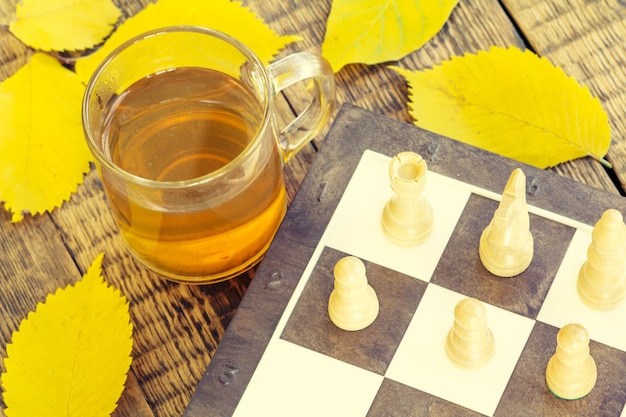 Piezas de ajedrez a bordo, taza de té y hojas amarillas de otoño sobre fondo de madera