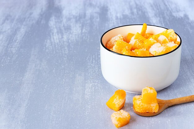 Piezas congeladas de calabaza en un recipiente blanco y una cuchara de madera en la mesa gris, espacio de copia