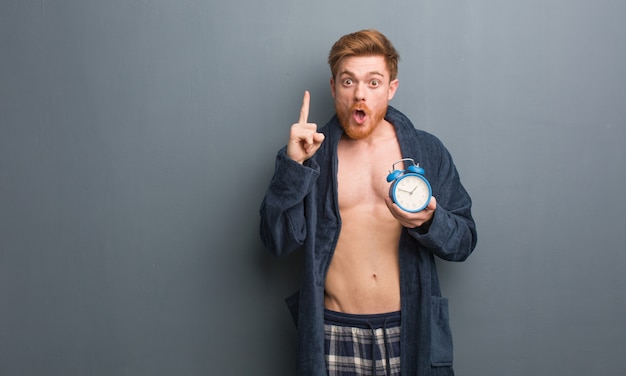 Pijama que lleva del hombre joven del pelirrojo que tiene una gran idea, concepto de creatividad. Él está sosteniendo un reloj de alarma.