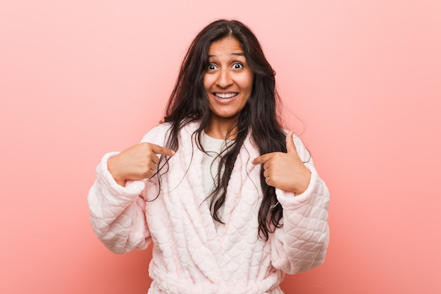 Pijama vestindo da mulher indiana nova surpreendido apontando nsi mesmo, sorrindo amplamente.