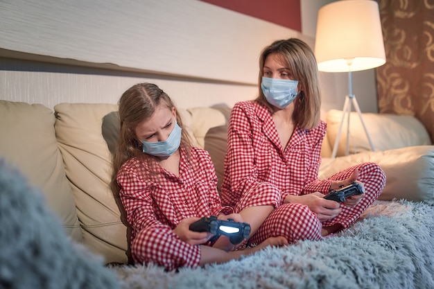 Pijamas vestindo da mulher e da moça e máscaras protetoras médicas que sentam-se no sofá na sala de visitas com quarentena do isolamento dos controladores do jogo de vídeo em casa auto, covid-19.