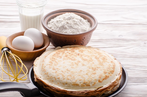 Pila de crepes franceses en sartén sobre mesa de cocina de madera con huevos de leche y harina a un lado