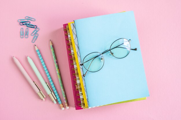 Pila de cuadernos con anteojos en la parte superior con grupos de clips, bolígrafos y lápices cerca sobre fondo rosa