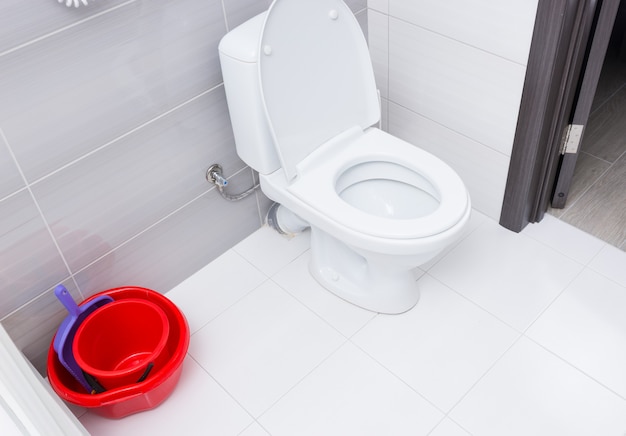 Pila de dos cubos de limpieza rojos con pala para recoger basura junto a abrir trabajado en el baño de la habitación del hotel con azulejos blancos y grises