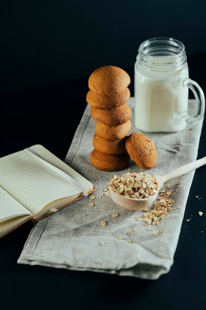 Pila de galletas de avena frescas con leche en tarro copos de avena cruda en una cuchara de madera grande cuaderno abierto sobre fondo negro Stilllife