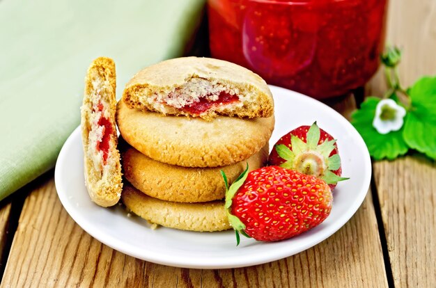 Una pila de galletas llenas de mermelada y fresas en un plato, un tarro de mermelada de fresa en el fondo de tablas de madera