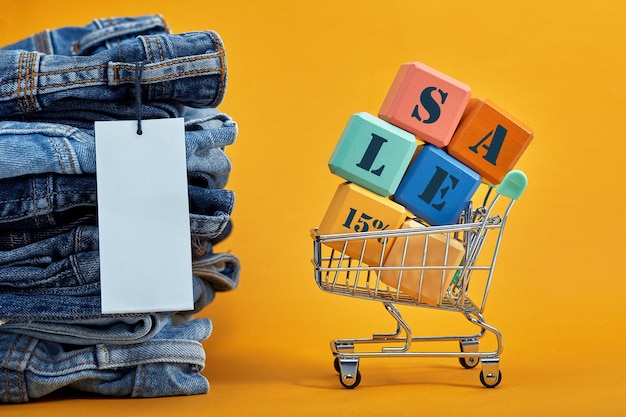 Una pila de jeans con una etiqueta en blanco sobre un fondo amarillo. Carro de compras con cubos multicolores. Ventas palabra escrita en cubos. Concepto de ventas. Montón de pantalones de mezclilla de moda con estilo.