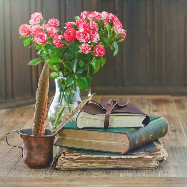 Pila de libros antiguos con ramo de flores rosas y una taza rústica con plumas se encuentran en una mesa de madera