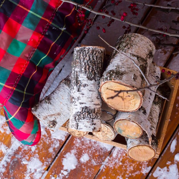 Una pila de madera de abedul cerca del sofá con una alfombra roja a cuadros en la terraza abierta de invierno cubierto de nieve