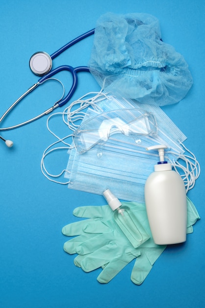 Pila de mascarillas médicas azules desechables, guantes de látex de goma, gafas protectoras, estetoscopio y alcohol antiséptico antiséptico para manos sobre fondo azul.