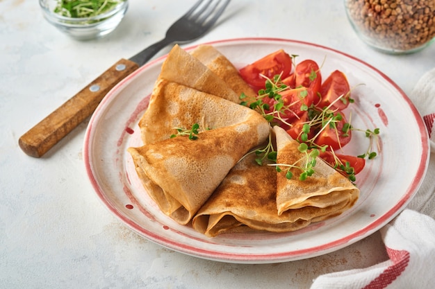 Pila de panqueques de crepes de harina de trigo sarraceno sin gluten con tomates cherry y microgreens de rúcula en un plato blanco, horneado saludable casero para el desayuno. Bosquejo. Copie el espacio.