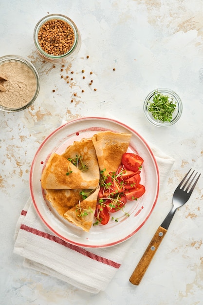 Pila de panqueques de crepes de harina de trigo sarraceno sin gluten con tomates cherry y microgreens de rúcula en un plato blanco, horneado saludable casero para el desayuno. Bosquejo. Copie el espacio.