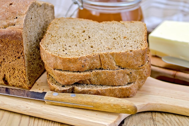 Una pila de rebanadas de pan casero de centeno con un cuchillo en un plato, servilleta, hogaza de pan, un tarro de miel, mantequilla sobre tabla de madera