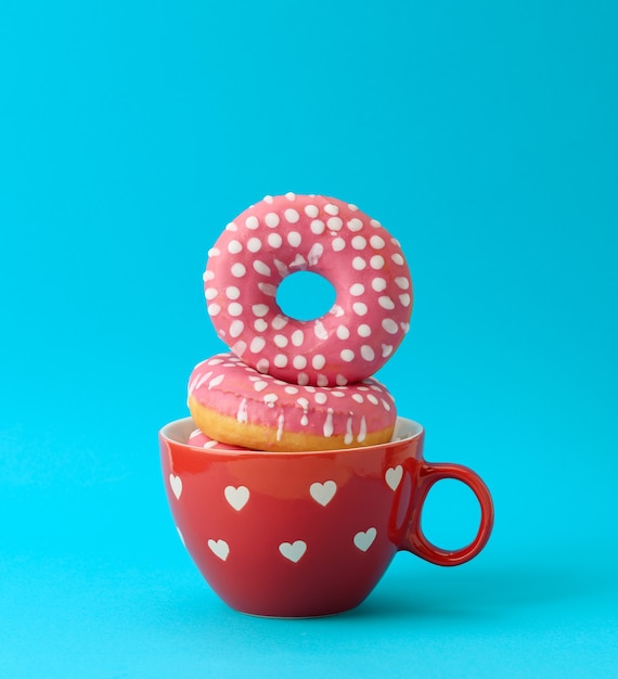 Una pila de rosquillas rosadas con glaseado en una taza de cerámica roja sobre una superficie azul, cerrar