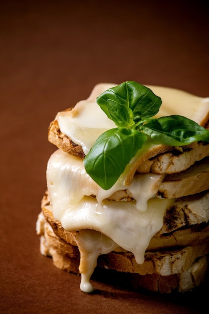 Pila de sándwiches prensados de queso derretido tostado con hojas de albahaca sobre una superficie de color marrón oscuro. De cerca