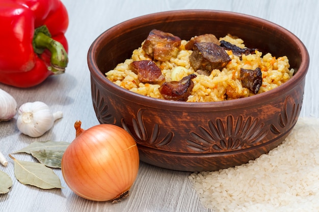 Pilaf de arroz en cuenco de barro con carne y verduras - cebolla, ajo, laurel, pimiento