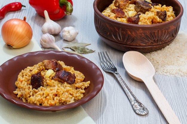 Pilaf de arroz en plato de barro y cuenco con carne y verduras: cebolla, ajo, pimiento rojo, pimiento. Cuchara de madera y tenedor de metal al lado de platos