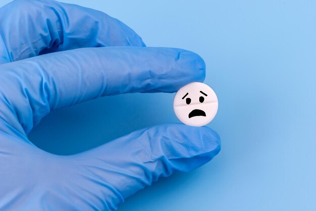 Una píldora con un emoticono triste en una mano con un guante médico sobre un fondo azul Atención médica