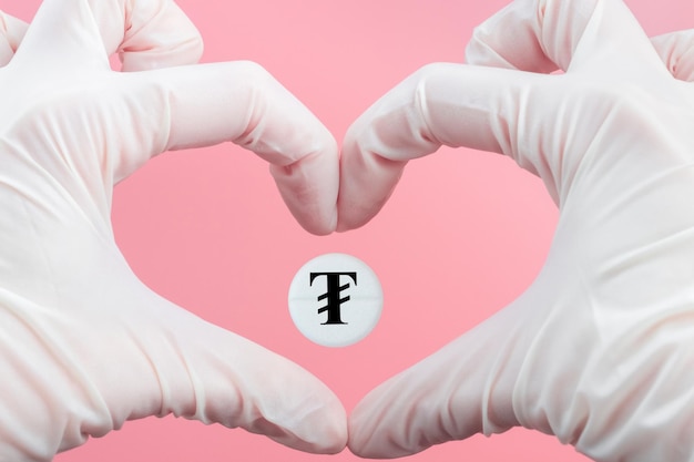 Foto una píldora con un signo de moneda tugrik dentro de un corazón hecho de manos con guantes médicos sobre un fondo rosa aumento del precio de los medicamentos en mongolia tratamiento costoso