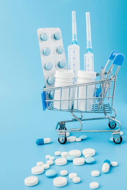 Píldoras e inyección médica en carrito de la compra sobre fondo azul. Idea creativa para el concepto de negocio de costo de atención médica, farmacia, seguro de salud y compañía farmacéutica. Copia espacio