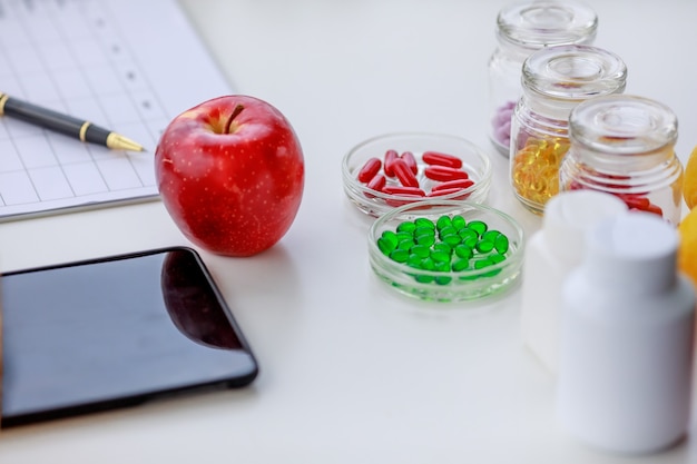 Píldoras de manzana, rojo, verde, amarillo en varios frascos, teléfono móvil y bolígrafo en la mesa de nutricionista con plan de alimentación semanal. Concepto de fotografía de vista superior de la mesa de nutrición.