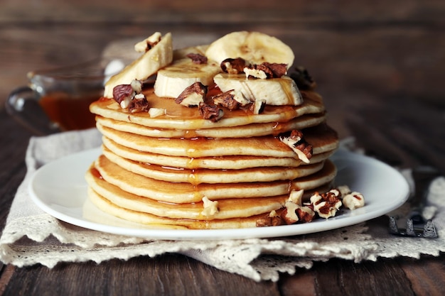 Pilha de deliciosas panquecas com nozes de mel de chocolate e fatias de banana no prato e guardanapo em fundo de madeira