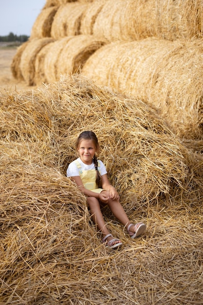 Pilha de feno no campo com uma menina sentada nele sorrindo com olhos pensativos manhosos olhando para longe segurando o feno na mão vestindo vestido de verão se divertindo longe da cidade no campo cheio de feno dourado