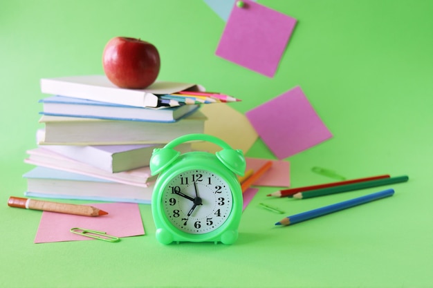 Pilha de livros didáticos, despertador, maçã e material escolar, fundo verde